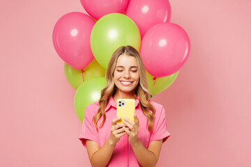 Young shocked fun woman wear shirt celebrating birthday holiday stand near bunch of colorful air balloons hold use mobile cell phone isolated on plain pastel light pink background. Lifestyle concept.