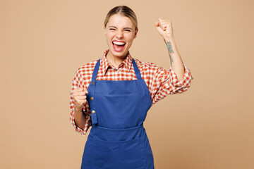 Wall Mural - Young woman barista bartender barman employee wears blue apron shirt work in coffee shop do winner gesture celebrate clenching fists isolated on plain beige background. Small business startup concept.