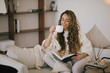 © polinaloves - Young woman reading book and drinking tea, sitting on sofa in a cozy living room.