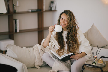 Young woman reading book and drinking tea, sitting on sofa in a cozy living room.