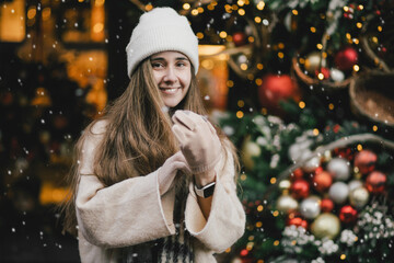 Young beautiful woman enjoying Christmas decorations, standing under snowfall on city street.