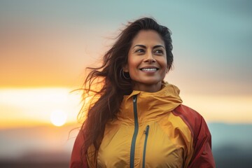 Wall Mural - Portrait of a smiling indian woman in her 40s wearing a functional windbreaker in front of vibrant sunset horizon