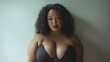 © Johannes - A woman with curly hair and a large bust stands in front of a white wall. Beautiful African American brunette plus size model wearing the black dress portrait. Body positivity and diversity