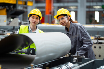 Two women wearing safety gear are looking at a large piece of metal. Scene is serious and focused, as the women are likely discussing the details of the metal piece or inspecting it for safety reasons