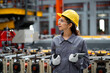 © KANGWANS - A woman wearing a yellow helmet and safety glasses stands in a factory. She is holding a tablet in her hand