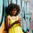 © CanYalicn - A cheerful woman in a yellow dress leans against a graffiti wall, smiling as she checks her phone. Vibrant colors and sunlight add warmth to her joyful, carefree moment.