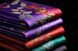 © Thanyarat - A stack of colorful Thai silk fabrics, each piece glowing with shades of purple, red, and emerald, with intricate floral and geometric patterns woven into the material.