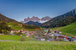 © Rafal Pawul - A picturesque Alpine village nestled in green hills with the stunning backdrop of the snow-capped Dolomite mountains. The serene landscape showcases beautiful homes and lush countryside under a clear