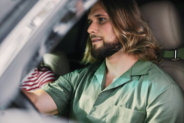 Wall Mural - Man with long hair and beard sitting in a car, wearing a green shirt Soft focus on the background