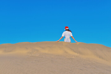 A girl in a dune wearing a Santa hat. Back view. The concept of a Christmas vacation in hot countries, at the sea. Dune near the sea in Maspalomas, Gran Canaria