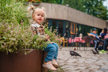 A cute toddler kid girl in a plaid shirt sits on the veranda of a European outdoor cafe in summer.