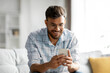© Home-stock - Young smiling hindu man holding mobile phone, typing sms or scrolling social media, sitting on couch in light living room interior, free space