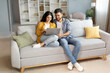 © Home-stock - Planning vacation. Happy young indian couple searching tour online, using laptop computer, sitting on couch in cozy living room interior at home, free space