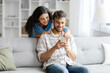 © Home-stock - Young hindu couple sharing social media on cellphone, happy indian spouses sitting on sofa, using smartphone gadget at home interior, woman embracing husband from back