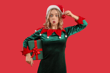 Sticker - Funny young woman dressed as elf with Christmas gift and apple on red background