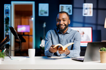 Wall Mural - Cheerful content creator in studio reviewing newly released hardcover book from publisher. African american reading hobbyist filming literature video essay, presenting new novel from author to readers