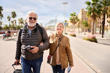 Wall Mural - Couple of happy Caucasian mature tourist walking looking at camera in street European city with their luggage. Pensioner people enjoying their holidays in winter sunny day