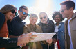 © CarlosBarquero - Group of diverse middle-aged tourist people standing looking at travel map in hands on street city on holidays smiling on sunny day. Mature friends gathered in community enjoying a trip. Low angle