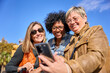 © CarlosBarquero - Group of diverse mature women smiling looking and using smartphone on a street. Middle-aged female friends searching a places on mobile and enjoying vacations on a autumn sunny day