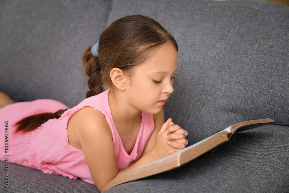 Cute little girl reading Bible on sofa at home