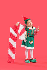 Sticker - Cute little girl in elf costume with candy cane pinata pointing at something on red background