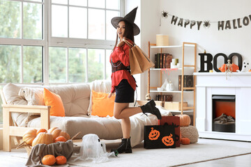 Sticker - Beautiful young happy Asian woman dressed as witch for Halloween with shopping bags in decorated living room