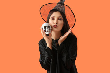 Poster - Beautiful young Asian woman dressed as witch for Halloween with human skull blowing kiss on orange background