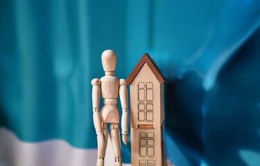 A wooden figurine stands near a miniature wooden house against a blue and white background.