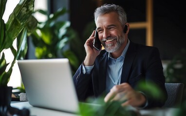 A professional man talks on the phone while working on his laptop in a green, plant-filled office environment.