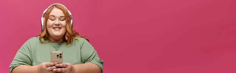 A woman in a green shirt listens to music while holding a phone in front of a pink background.