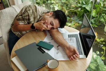 High angle view of young man lying on his workplace with laptop and documents, he tired of his work