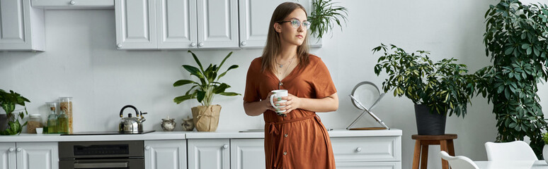 A woman in an orange dress holds a coffee mug in a white kitchen.