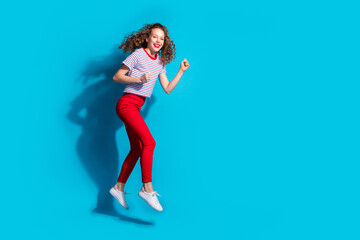 Wall Mural - Cheerful young woman jumping joyfully in vibrant red pants and striped shirt against blue background, exuding high energy positive emotion in studio setting