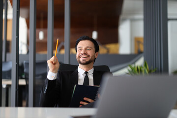 Wall Mural - A man in a suit is sitting at a desk with a pencil in his hand and a book in front of him. He is smiling and he is in a positive mood. Concept of productivity and focus