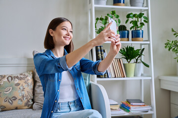 Beautiful happy young woman taking selfie photo on smartphone in home interior