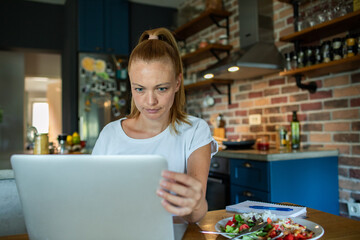 Wall Mural - Smiling woman eating salad while video chatting from home kitchen