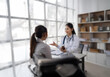 © YiuCheung - A female doctor in a white coat holds a medicine bottle and speaks with a patient sitting across from her at a desk.