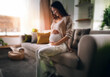 © YiuCheung - A pregnant woman sits on a couch, cradling her stomach and smiling softly, bathed in warm, natural light.