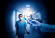 © YiuCheung - A gloved hand holds a syringe with blue liquid in a medical setting, another person in scrubs walks in the background.