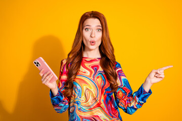 Wall Mural - Photo of attractive young woman hold device amazed point empty space dressed stylish colorful clothes isolated on orange color background