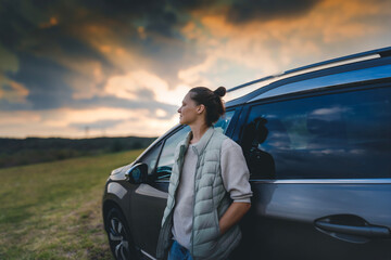 Wall Mural - Young female driver standing by car enjoying sunset and nature in field during road trip