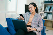 © Parichat - Young asian businesswoman sitting on blue sofa working from home, using smartphone and laptop computer, smiling and looking at mobile phone screen