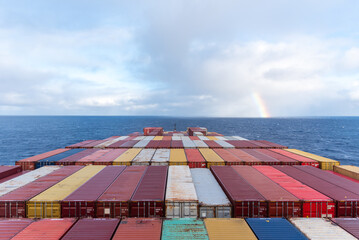 The cargo ship sails into the horizon, its deck stacked with containers. Clouds and rainbow pops up on the horizon. 