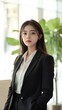 © EarthWalker - Portrait of a young beautiful Asian businesswoman. Confident female business manager in black suit looking into camera in modern office with a green indoor plant.