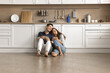 © fizkes - Happy couple of homeowners sitting on floor close together in new spacious kitchen interior, drinking tea with white furniture, utensils in background, looking away, smiling, laughing