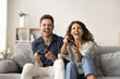 © fizkes - Joyful happy couple excited with video game, virtual entertainment, holding gamepads, pushing buttons of game controllers, shouting, laughing, pushing buttons, sitting on home sofa, front view