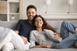 © fizkes - Happy pretty young wife and handsome husband enjoying romantic relationship, leisure on home couch together, hugging with love, affection, looking at camera with toothy smiles