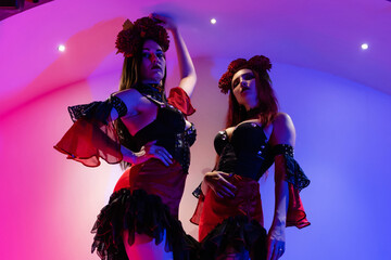 Wall Mural - Two beautiful girls in Mexican dresses and a herbarium on their heads. They stand in the semi-darkness, illuminated by blue and violet light. Halloween concept, nightclub, masquerade