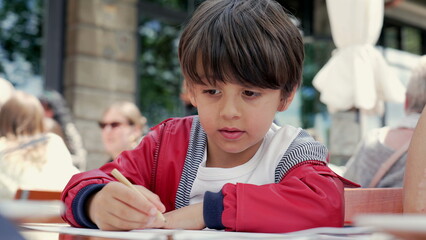 Young boy in a red jacket focused on drawing with a pencil at an outdoor restaurant, with people in the blurred background enjoying the sunny day