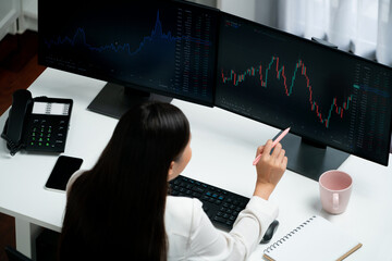 Young beautiful Asian businesswoman looking on pc to analyze profitable of exchange stock market graph at modern office. Concept of investing high profit in wealth security in finance. Stratagem.